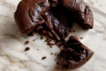 Brownie que deu errado com centro cru e massa úmida escorrendo, aberto sobre bancada de mármore com pedaço ao lado.