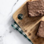 Quadrados de brownie recém-assados dispostos sobre uma tábua de madeira em uma bancada de mármore branco, com um pano xadrez e uma faca ao lado.