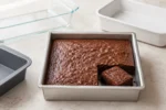Brownie assado em forma quadrada de alumínio claro, acompanhado por formas de vidro, cerâmica e silicone.