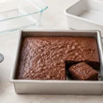 Brownie assado em forma quadrada de alumínio claro, acompanhado por formas de vidro, cerâmica e silicone.