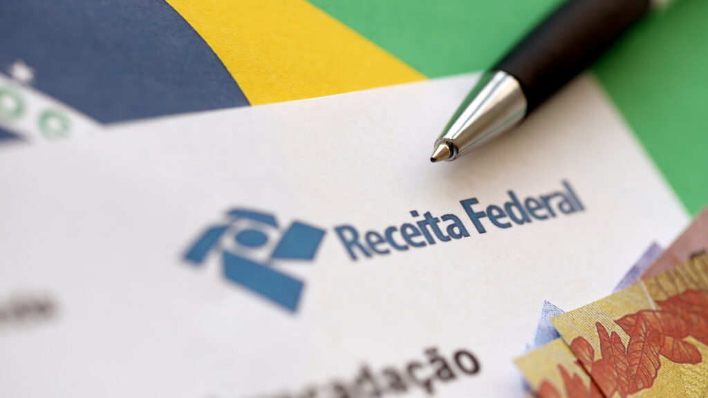 das-documento-arrecadacao Folha impressa com o logotipo da Receita Federal ao lado de uma caneta preta e prateada, parcialmente coberta por cédulas de real sobre uma superfície com as cores da bandeira do Brasil