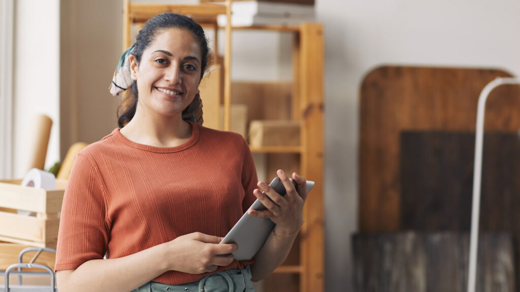 mei-micro-empreendedor-individual Mulher sorridente de cabelos presos e blusa laranja segurando um tablet em um ambiente de trabalho com prateleiras de madeira ao fundo