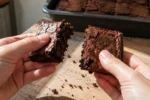 Mãos partindo um brownie seco e firme ao meio, com migalhas sobre uma tábua
