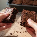 Mãos partindo um brownie seco e firme ao meio, com migalhas sobre uma tábua