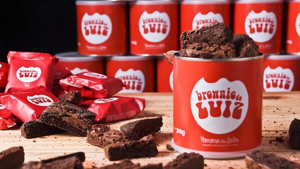 Brownies do Luiz em embalagem vermelha, com latas e unidades embaladas sobre mesa de madeira.