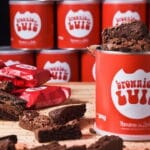 Brownies do Luiz em embalagem vermelha, com latas e unidades embaladas sobre mesa de madeira.