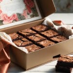 Caixa dourada aberta com brownies organizados, papel decorado e laço de cetim sobre mesa rústica branca