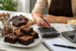 Brownies na bancada com calculadora, caderno e ingredientes, mostrando preço do brownie para vender com lucro.