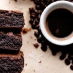 Pilha de três brownies de café ao lado de uma xícara de café preto e grãos de café espalhados sobre a mesa.