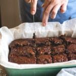 Close-up de mãos cortando um brownie assado em pedaços quadrados em uma forma forrada com papel manteiga.