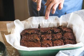 Close-up de mãos cortando um brownie assado em pedaços quadrados em uma forma forrada com papel manteiga.