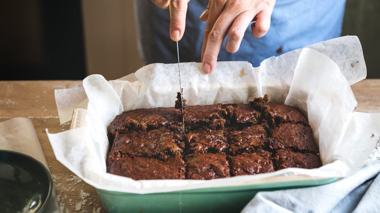 Close-up de mãos cortando um brownie assado em pedaços quadrados em uma forma forrada com papel manteiga.