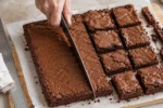 Mão cortando brownie em quadrados ao lado de uma régua e aparas sobre papel-manteiga