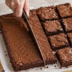 Mão cortando brownie em quadrados ao lado de uma régua e aparas sobre papel-manteiga