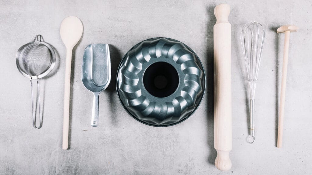 Utensílios de cozinha como peneira, colher de pau, forma de bolo, rolo de massa e batedor dispostos sobre uma superfície cinza.