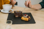 Brownie gourmet sobre prato preto ao lado de xícara de café.