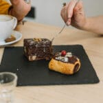 Brownie gourmet sobre prato preto ao lado de xícara de café.