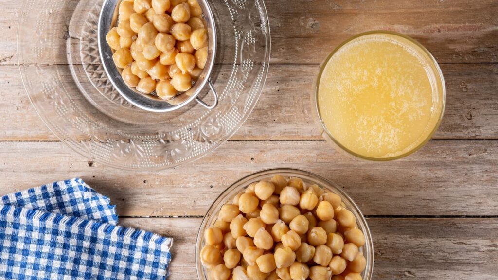 aquafaba Grãos de grão-de-bico cozidos escorrendo em peneira sobre prato de vidro, ao lado de tigela com líquido amarelo translúcido de aquafaba, ingrediente usado para substituir claras de ovo e criar casquinha brilhante em brownie vegano