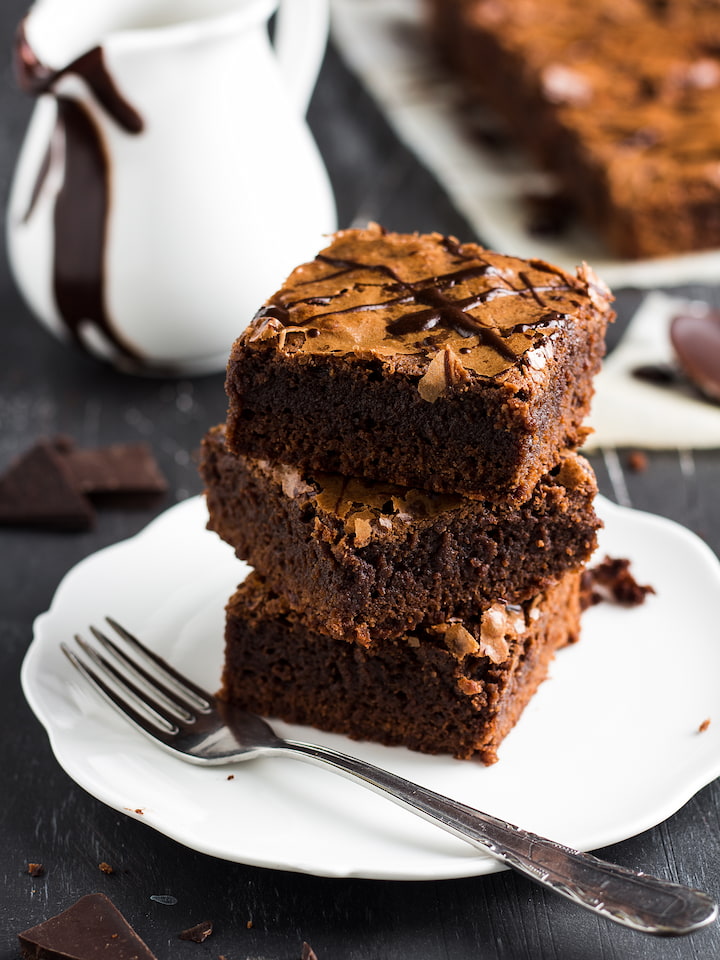 brownie-vegano Três pedaços de brownie vegano empilhados em prato branco, com casquinha crocante e rachada na superfície, regados com calda de chocolate, garfo de metal ao lado, jarra branca com calda ao fundo sobre mesa escura
