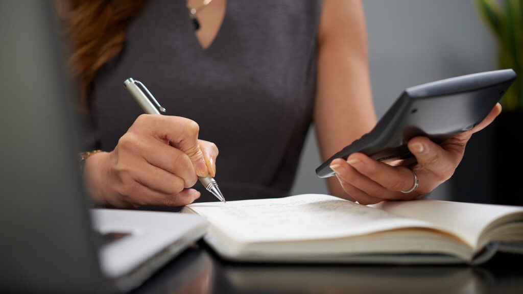 ferramentas-gestao-financeira Mãos de confeiteira fazendo o controle financeiro do negócio, calculando custos e anotando em um caderno ao lado de um notebook.