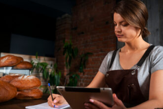 Confeiteira de avental faz a gestão financeira de sua confeitaria usando um tablet e anotando em um caderno, em um balcão de madeira com pães ao fundo.