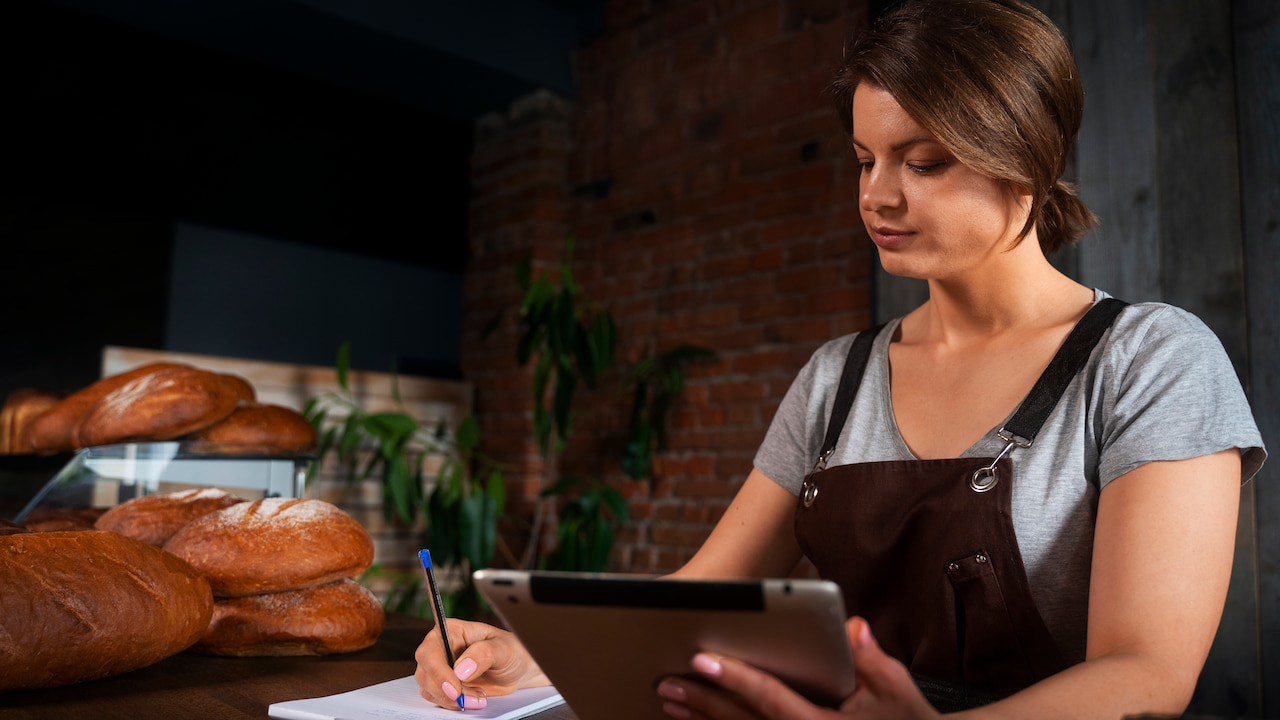 Confeiteira de avental faz a gestão financeira de sua confeitaria usando um tablet e anotando em um caderno, em um balcão de madeira com pães ao fundo.