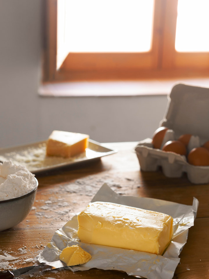 mesa-com-manteiga tablete de manteiga parcialmente cortado sobre papel manteiga em mesa de madeira, com farinha e ovos ao fundo, representando preparo de ingredientes para brownie