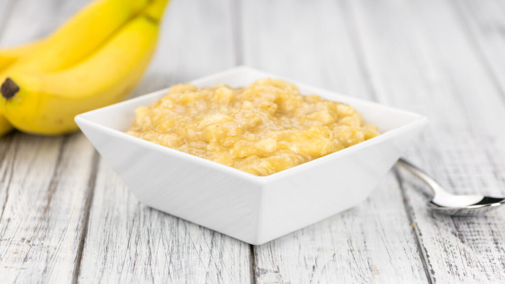 pure-de-banana Purê de banana amarelo cremoso em tigela branca quadrada sobre mesa de madeira clara, com bananas maduras ao fundo e colher de metal ao lado, ingrediente usado como substituto do ovo para brownie vegano macio e úmido