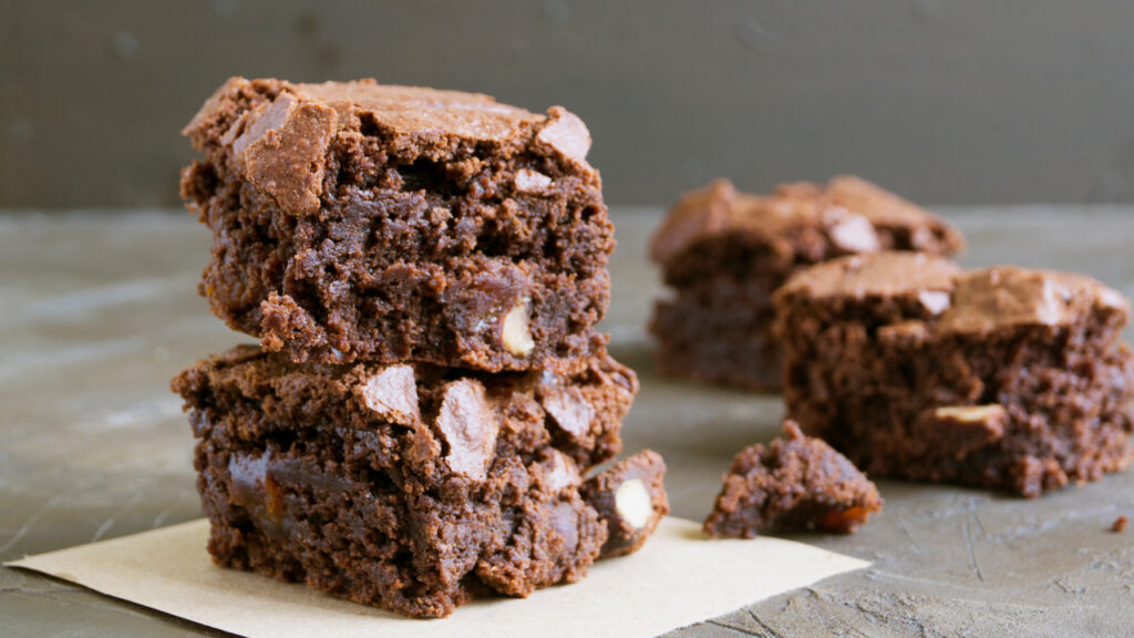Dois pedaços de brownie low carb empilhados, com massa escura e úmida, textura quebradiça na superfície, sobre papel manteiga, com outros pedaços desfocados ao fundo