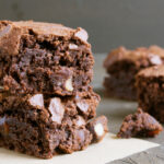 Dois pedaços de brownie low carb empilhados, com massa escura e úmida, textura quebradiça na superfície, sobre papel manteiga, com outros pedaços desfocados ao fundo