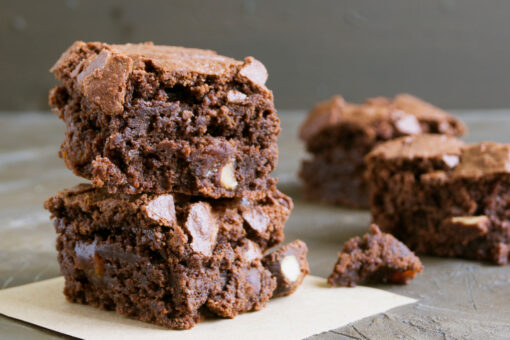 Dois pedaços de brownie low carb empilhados, com massa escura e úmida, textura quebradiça na superfície, sobre papel manteiga, com outros pedaços desfocados ao fundo