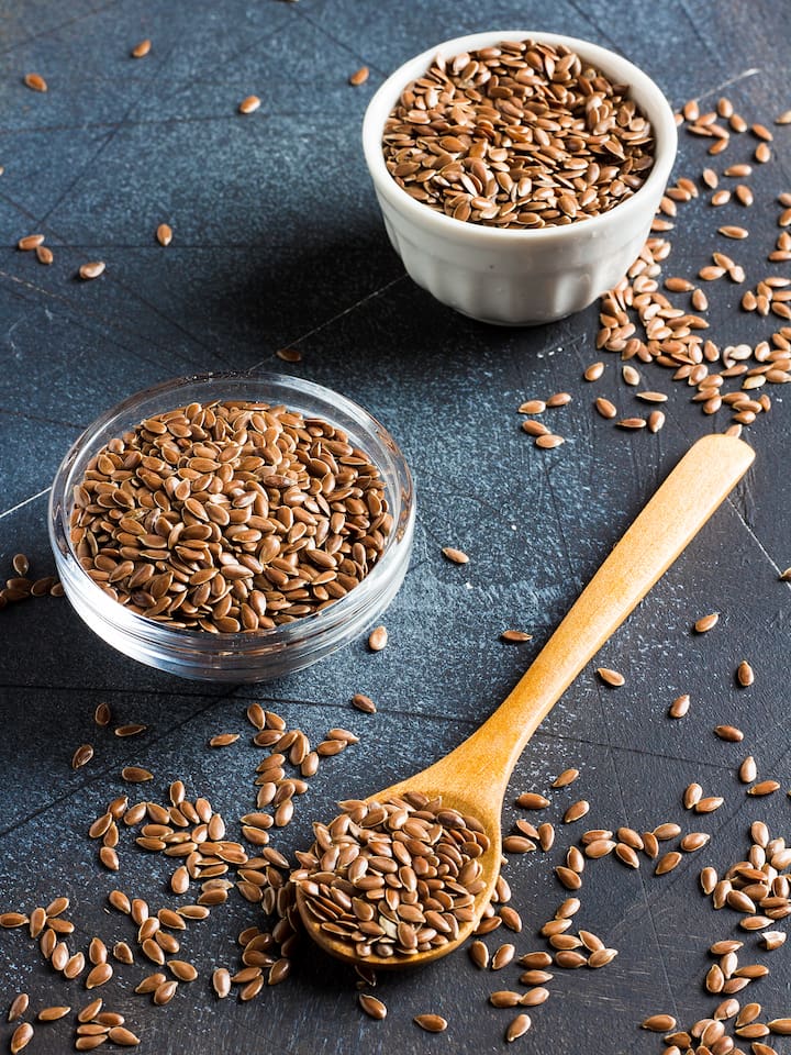 sementes-de-linhaca Sementes de linhaça marrons inteiras espalhadas sobre superfície escura, dentro de colher de madeira, tigela de vidro e ramequim branco, ingrediente usado para formar gel viscoso substituto do ovo em brownies veganos densos e úmidos