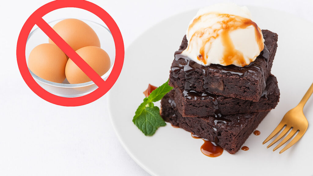Dois pedaços de brownie vegano de chocolate escuro empilhados, cobertos com calda brilhante e bola de sorvete branco, decorados com hortelã, ao lado de garfo dourado, com símbolo de proibido sobre tigela de ovos, representando receita sem ovos
