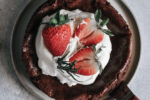 Brownie de frigideira com bordas crocantes coberto com chantilly, morangos frescos inteiros e cortados ao meio, e ramos de alecrim, servido em prato sobre superfície cinza