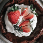 Brownie de frigideira com bordas crocantes coberto com chantilly, morangos frescos inteiros e cortados ao meio, e ramos de alecrim, servido em prato sobre superfície cinza