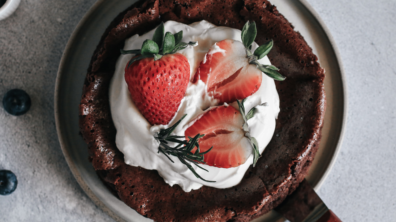 Brownie de frigideira com bordas crocantes coberto com chantilly, morangos frescos inteiros e cortados ao meio, e ramos de alecrim, servido em prato sobre superfície cinza