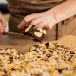 Pessoa usando espátula para cortar pedaço de brownie caseiro coberto com castanhas laminadas, sobre papel manteiga em bancada de cozinha doméstica