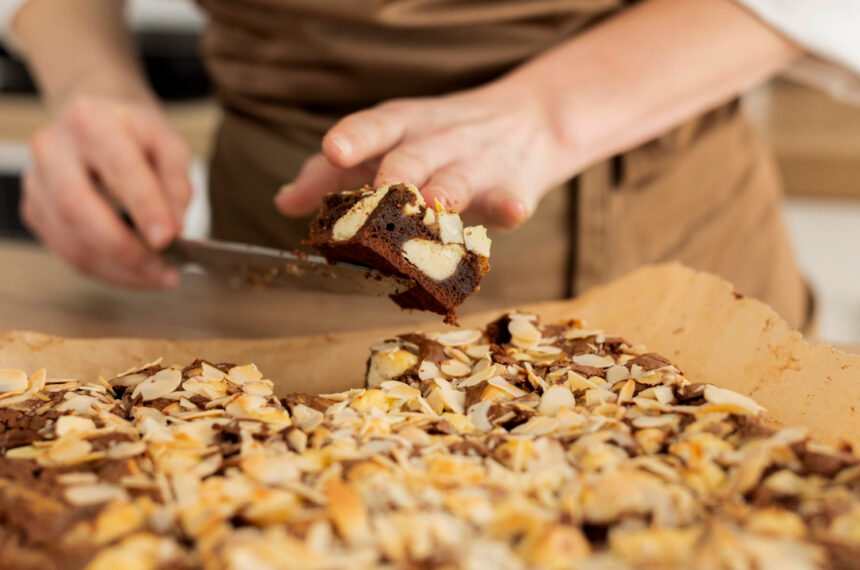 Pessoa usando espátula para cortar pedaço de brownie caseiro coberto com castanhas laminadas, sobre papel manteiga em bancada de cozinha doméstica