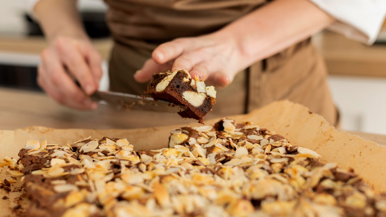 Pessoa usando espátula para cortar pedaço de brownie caseiro coberto com castanhas laminadas, sobre papel manteiga em bancada de cozinha doméstica