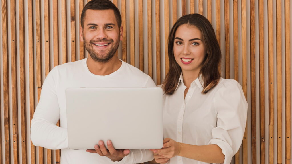 conciliar-emprego-clt-com-empreendedorismo Homem e mulher sorrindo, vestindo roupas brancas, segurando um notebook juntos em frente a uma parede de ripas de madeira vertical