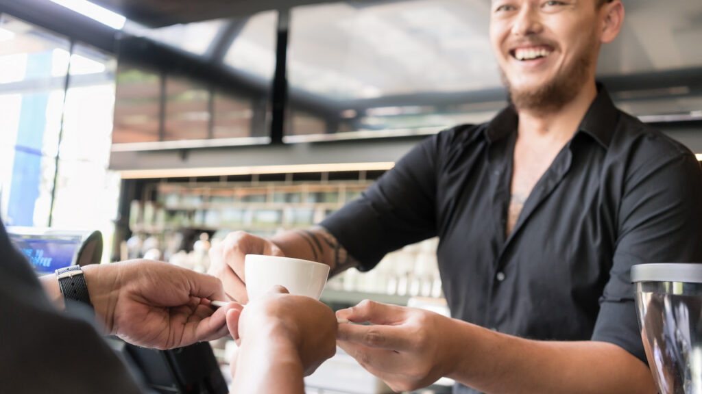 parceria-em-cafeterias Homem sorridente com camisa preta servindo uma xícara de café branco para um cliente em uma cafeteria moderna, simbolizando parceria comercial para venda de produtos como brownies