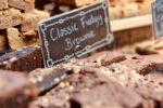 Brownies de chocolate com pedaços de nozes organizados sobre uma bancada de feira com placa escrita “Classic Fudgy Brownie” ao fundo, ao lado de outros tipos de brownies expostos para venda