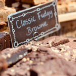 Brownies de chocolate com pedaços de nozes organizados sobre uma bancada de feira com placa escrita “Classic Fudgy Brownie” ao fundo, ao lado de outros tipos de brownies expostos para venda
