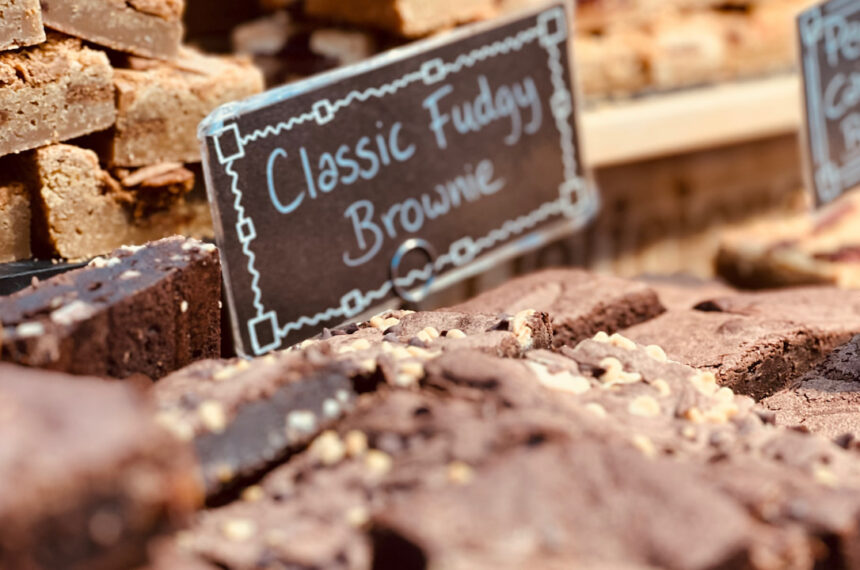Brownies de chocolate com pedaços de nozes organizados sobre uma bancada de feira com placa escrita “Classic Fudgy Brownie” ao fundo, ao lado de outros tipos de brownies expostos para venda