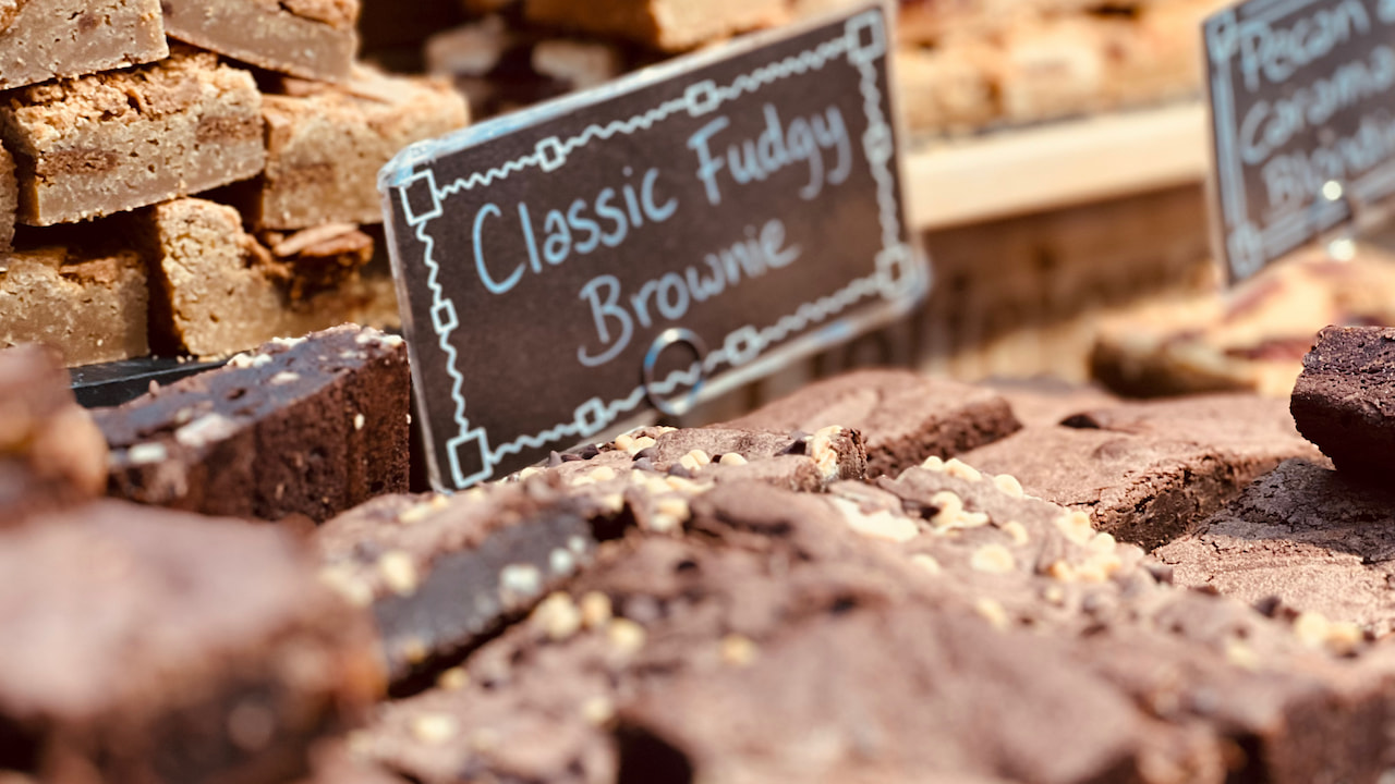 Brownies de chocolate com pedaços de nozes organizados sobre uma bancada de feira com placa escrita “Classic Fudgy Brownie” ao fundo, ao lado de outros tipos de brownies expostos para venda