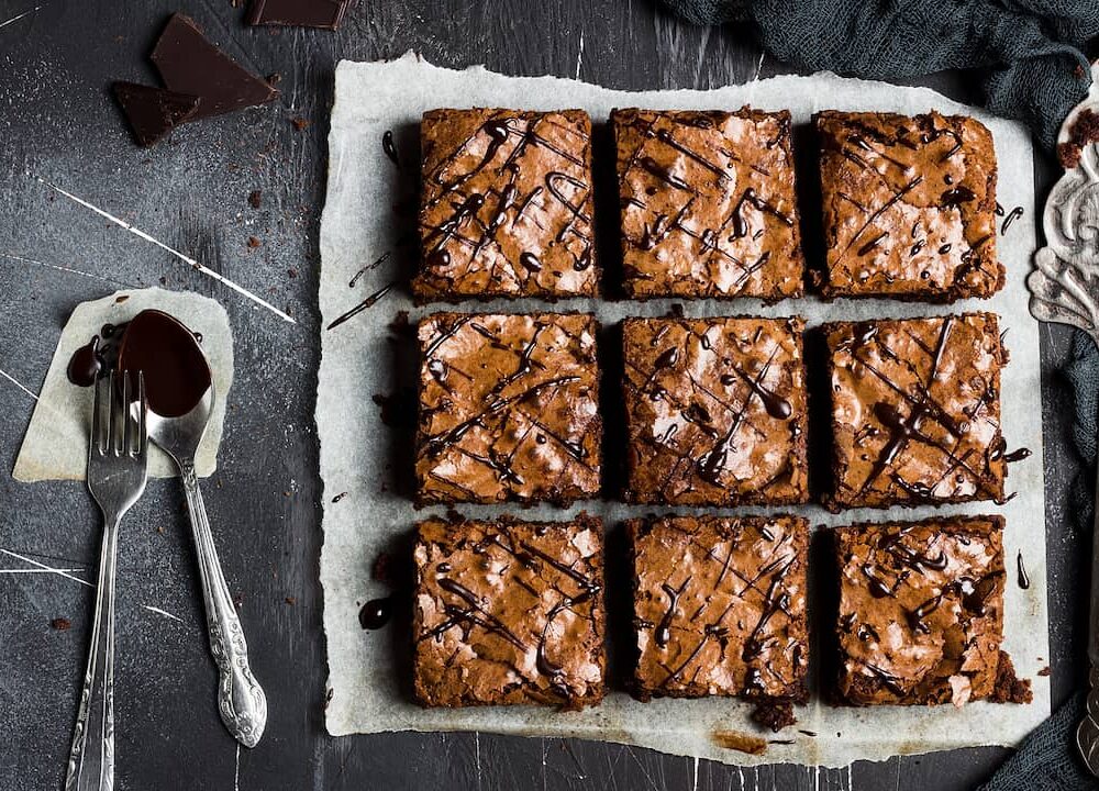Nove quadrados de brownie com casquinha craquelada e fios de chocolate sobre papel manteiga, ao lado de garfo e colher com calda em fundo escuro; bordas firmes e centro úmido, ilustrando o ajuste do tempo de forno do brownie.