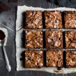 Nove quadrados de brownie com casquinha craquelada e fios de chocolate sobre papel manteiga, ao lado de garfo e colher com calda em fundo escuro; bordas firmes e centro úmido, ilustrando o ajuste do tempo de forno do brownie.