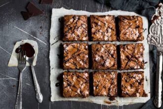 Nove quadrados de brownie com casquinha craquelada e fios de chocolate sobre papel manteiga, ao lado de garfo e colher com calda em fundo escuro; bordas firmes e centro úmido, ilustrando o ajuste do tempo de forno do brownie.