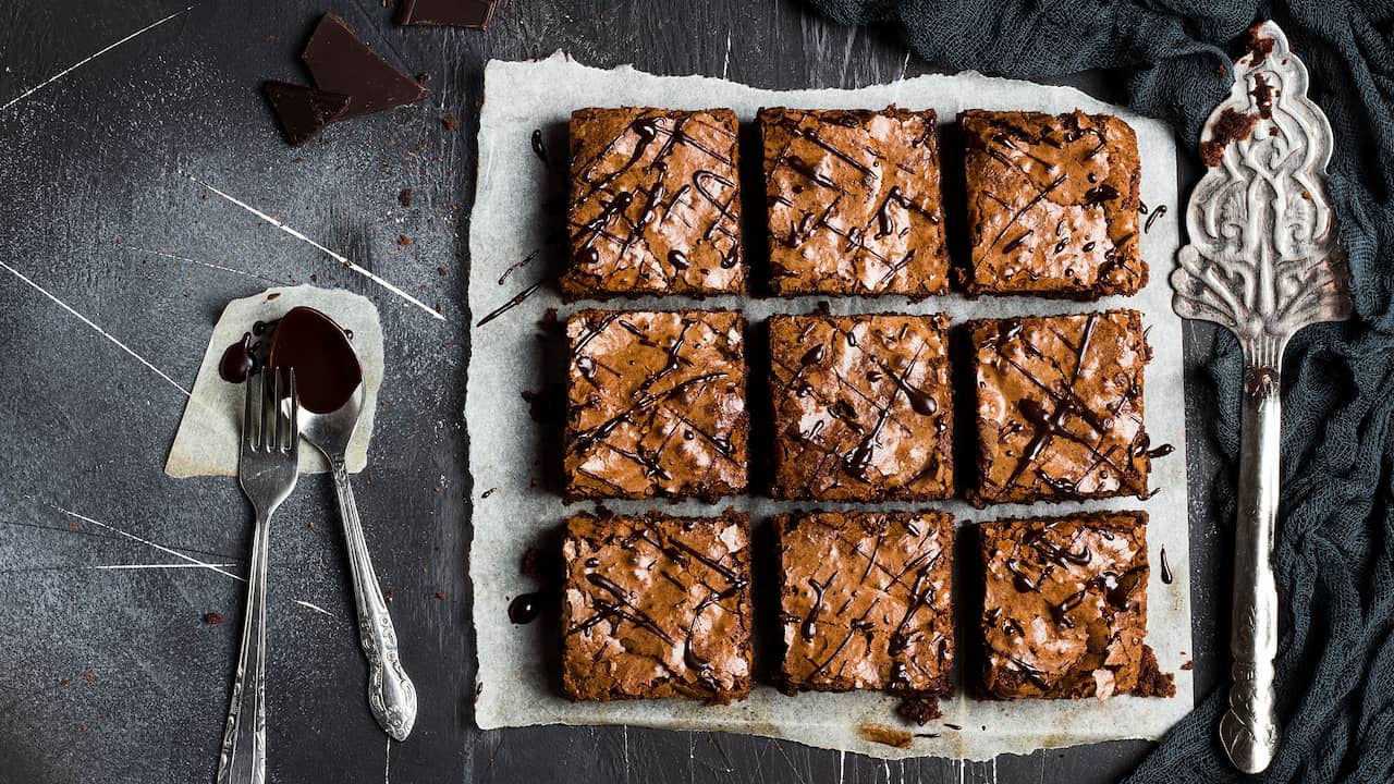 Nove quadrados de brownie com casquinha craquelada e fios de chocolate sobre papel manteiga, ao lado de garfo e colher com calda em fundo escuro; bordas firmes e centro úmido, ilustrando o ajuste do tempo de forno do brownie.
