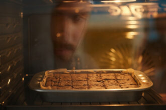 Brownie de chocolate assando em forno doméstico, cortado em quadrados, com crosta brilhante e rachada, observado por uma pessoa através do vidro